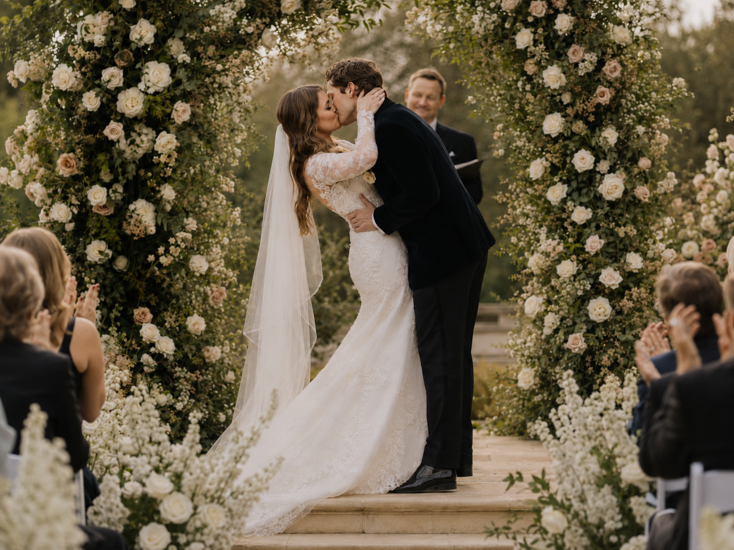 Newlyweds sharing their first kiss under a floral arch as guests applaud